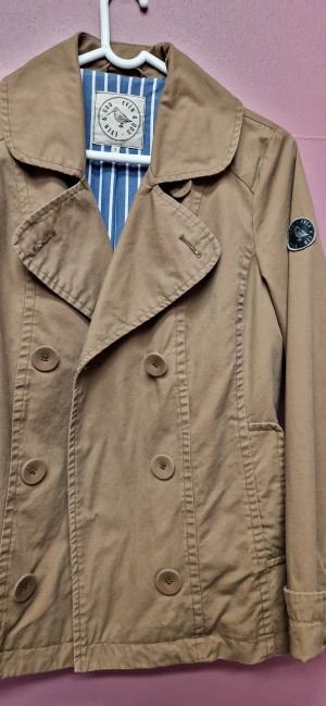 Beige skepparkavaj från Even & Odd - Snygg beige skepparkavaj från Even & Odd med klassisk dubbelknäppning och bred krage. Jackan har ett coolt tygmärke på ärmen och är tillverkad i bomull. Insidan har blåvita ränder som ger extra stilpoäng. Perfekt för dig som gillar tidlös look.