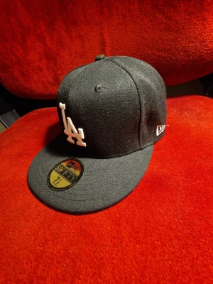 LA keps  - Säljer en svart New Era 59FIFTY keps med LA-logga i vitt framtill. Klassisk flat brim och broderad logga på sidan. Kepsen är tillverkad i bomull och har en stilren look som passar perfekt till streetwear. Storlek 7 1/8 (56,8 cm).