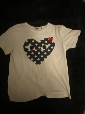 Vit Comme des Garçons Play t-shirt - Säljer en vit t-shirt från Comme des Garçons Play med ikoniskt hjärtmotiv på bröstet. Stort mörkblått hjärta med vita prickar och ögon, samt ett litet rött hjärta. Klassisk rund hals och korta ärmar. Materialet är mjuk bomull, perfekt för en clean streetstyle.