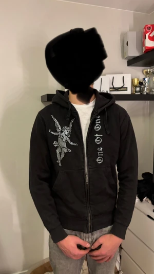 Svart hoodie med ängeltryck One Of One - Svart hoodie från One Of One med dragkedja och huva. På framsidan finns ett stort tryck av en ängel i silver och texten 'One Of One' i glittriga detaljer. Tröjan har långa ärmar, ribbade muddar och en klassisk loose passform. Perfekt för dig som gillar streetwear.