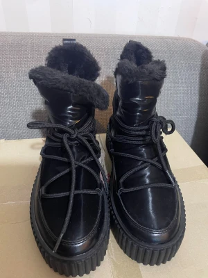 Svarta moon boots med fuskpäls - Säljer ett par svarta moon boots med glansig yta, fuskpälsfoder och kraftig räfflad sula. Skorna har snörning runt om och är perfekta för kalla dagar. De är riktigt chunky och har en modern vibe som sticker ut.