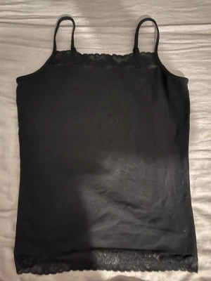 Svart spetskantad linnetopp Lindex - Svart linnetopp från Lindex med smala axelband och fina spetsdetaljer längs både halsringning och nederkant. Toppens material är mjukt och stretchigt, perfekt för en enkel och stilren look. Passar dig som gillar minimalistisk stil med en touch av spets.