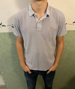 Ljusgrå pikétröja från Ralph Lauren - Snygg ljusgrå pikétröja från Ralph Lauren med klassisk krage och två knappar framtill. Tröjan har korta ärmar och den ikoniska orangea Polo-loggan broderad på bröstet. Tillverkad i 100% bomull för skön och luftig känsla.