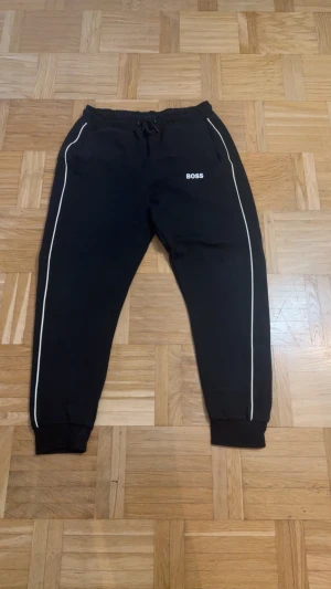 Svarta mjukisbyxor från BOSS - Svarta mjukisbyxor från BOSS med vita ränder längs benen och logga på vänster sida. Byxorna har resår i midjan med snörning och muddar vid bensluten. Perfekta för en chill och sportig look.