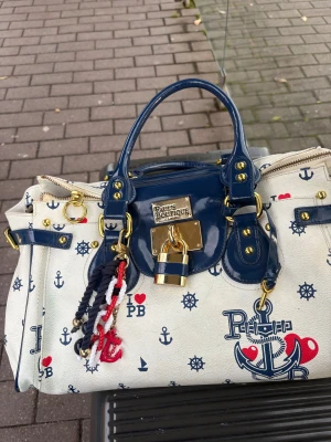 Chloe Paddington  väska - Snygg handväska från Pauls Boutique med marinblå detaljer och guldfärgade metalldelar. Väskan är vit med nautiskt mönster av ankare, hjul och hjärtan i blått och rött. Dekorativt hängande rep och lås framtill. Perfekt för dig som letar efter en chloe Paddington liknade väska