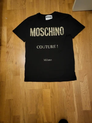 Svart Moschino Couture t-shirt - Svart t-shirt från Moschino med stort guldigt tryck 'MOSCHINO COUTURE! Milano' på bröstet. Klassisk passform med rund hals och korta ärmar. Tillverkad i 100% bomull för en skön och mjuk känsla. Perfekt statement-plagg för dig som gillar exklusiva märken.