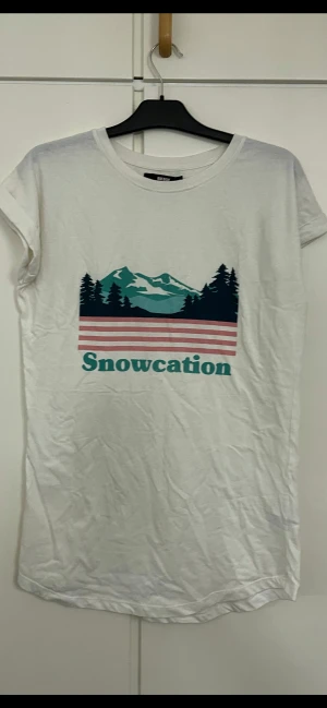 Vit Snowcation t-shirt från Bik Bok - Vit t-shirt från Bik Bok i storlek XS med coolt tryck framtill som visar berg, skog och rosa ränder samt texten 'Snowcation'. Rund hals och korta ärmar, tillverkad i mjuk bomull. Perfekt för dig som gillar grafiska prints och chill vibes.