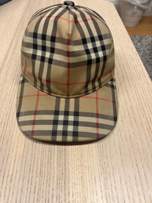 Burberry rutig keps i beige och svart - Säljer en snygg Burberry keps med klassiskt rutigt mönster i beige, svart och röda linjer. Kepsen har justerbar snapback och en svart Burberry London England patch bak. Materialet är bomull och polyester, perfekt för dig som vill ha en ikonisk streetstyle-accessoar.