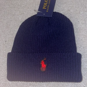 Mörkblå och svart mössa Polo Ralph Lauren - Två ribbstickade mössor från Polo Ralph Lauren, en i mörkblått och en i svart. Båda har uppvikt kant och den klassiska röda broderade polospelaren framtill. Perfekt accessoar för kyliga dagar med stilren och tidlös design.