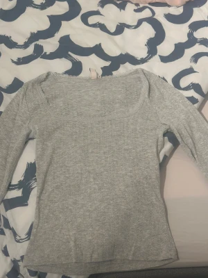 Ljusgrå ribbad långärmad topp - Säljer en ljusgrå långärmad topp med ribbad struktur och bred, rundad halsringning. Toppen är mjuk och stretchig, perfekt för en chill och enkel look. Passar dig som gillar basic med lite extra detalj i tyget.