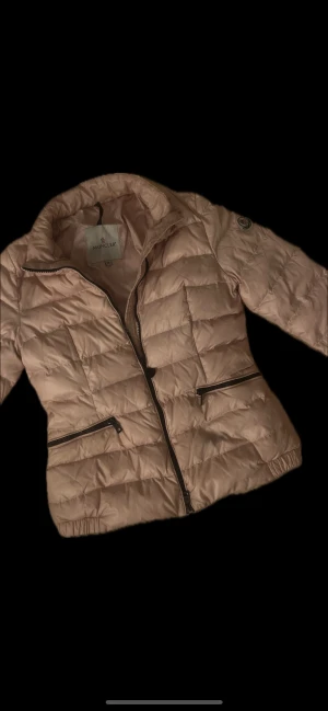 Ljusrosa Moncler pufferjacka - Moncler lättviktsjacka – rosa, dam  Stilren och exklusiv lättviktsjacka i en mjukt rosa ton. Jackan har klassisk Moncler-design med quiltat mönster, hög krage och märkespatch på ärmen. Lätt, varm och perfekt för vår, höst eller mildare vinterdagar.  Skick: Mycket bra begagnat skick – inga defekter, fläckar eller slitningar. Detaljer: 	•	Två blixtlåsfickor 	•	Hel dragkedja fram 	•	Elastiska ärmslut nedtill 	•	Lätt och bekväm dun-/syntetfyllning (baserat på modelltyp) 	•	Ikoniskt Moncler-emblem på