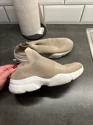 Beige stickade sneakers med platt sula - Säljer ett par beige sneakers med stickad ovandel och vit, chunky platt sula. Skorna har en strumpliknande design utan snörning . Perfekt för dig som gillar stilrena och bekväma skor. Storlek 37-38. Använd ett fåtal gånger.