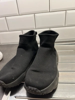 Svarta sock-sneakers med platt sula - Svarta sneakers med stickad ovandel som sitter som en strumpa runt foten. Skorna har en chunky platt sula i svart och en rund tå. Perfekt för dig som gillar minimalistisk och modern stil. Materialet är syntet och stickat tyg. Storlek 38. Dom är använd 5-7 gånger. 