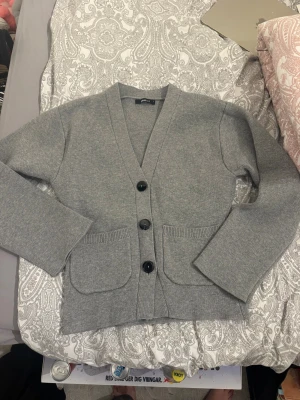 Grå kofta från Gina Tricot - Mysig grå kofta från Gina Tricot i storlek S. Koftan har v-ringning, tre svarta knappar framtill och två stora fickor. Materialet är stickat och känns mjukt mot huden. Perfekt att slänga på sig när det är lite kyligt ute.