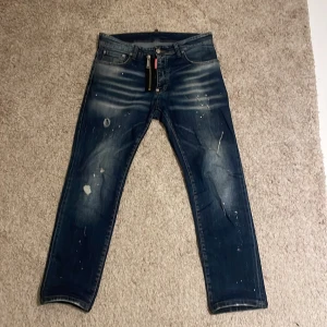 Dsquared2 jeans - Säljer ett par mörkblå jeans från Dsquared2 i storlek 50. Jeansen har coola slitningar, färgstänk och en snygg tvättad look. Klassisk femficksmodell med Dsquared2-logga bak. Perfekta för dig som gillar streetstyle och vill sticka ut.