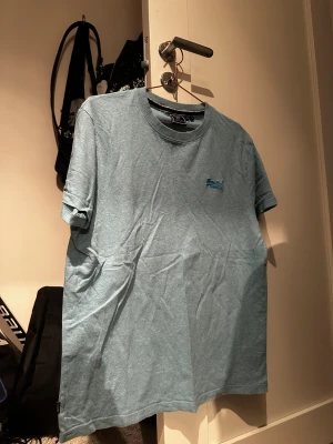 Blå t-shirt från Superdry i bomull - Snygg ljusblå t-shirt från Superdry i ekologisk bomull. Klassisk rund hals och korta ärmar, med diskret Superdry-logga på bröstet. Perfekt basic att ha i garderoben, enkel att matcha med jeans eller shorts.