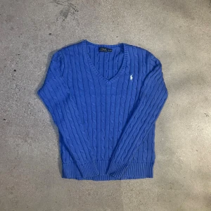 Blå kabelstickad tröja Polo Ralph Lauren - Kabelstickad blå tröja från Polo Ralph Lauren med v-ringning och broderad vit logga på bröstet. Tröjan har långa ärmar, ribbade muddar och är gjord i mjuk bomull. Storlek herr large.