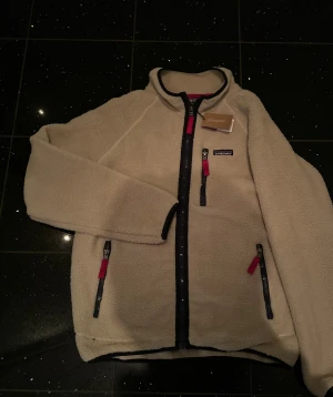 Beige fleecejacka från Patagonia L - Säljer en beige fleecejacka från Patagonia i storlek L men passar m för lite oversized. Jackan har mörkblå dragkedjor med röda detaljer, hög krage och tre fickor med dragkedja. Mjuk teddystruktur och klassisk Patagonia-logga på bröstet. Perfekt för dig som gillar outdoor och chill stil.