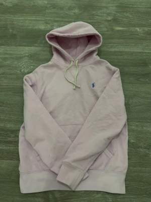 Ljusrosa hoodie från Polo Ralph Lauren - Säljer en ljusrosa hoodie från Polo Ralph Lauren med klassisk blå logga på bröstet. Tröjan har huva med snörning och en stor magficka. Materialet är mjukt och består av 60% bomull och 40% polyester. Perfekt för chill dagar och snygg till jeans.