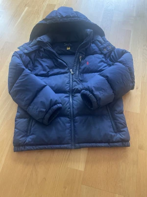 Mörkblå pufferjacka från Polo Ralph Lauren - Snygg mörkblå pufferjacka från Polo Ralph Lauren med huva och två sidofickor med dragkedja. Jackan har det klassiska röda Polo-logot broderat på bröstet och RL-detalj på dragkedjan. Perfekt för kalla dagar. Perfekt julklapp till någon du tycker om eller som en välförtjänt present till dig själv 