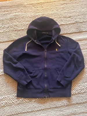 Marinblå hoodie från Polo Ralph Lauren - Snygg marinblå hoodie från Polo Ralph Lauren med gul broderad logga på bröstet. Tröjan har huva med vita snören, dragkedja framtill och två fickor. Tillverkad i mjuk bomull och har långa ärmar med ribbade muddar. Perfekt för chill dagar. Saknar dragaren på dragkedjan (bild) där av de låga priset!