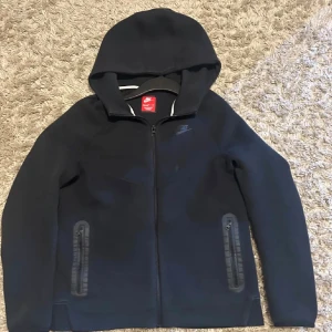 Svart Nike vindjacka med huva XL - Säljer en svart vindjacka från Nike i storlek XL. Jackan har huva, hel dragkedja framtill och två fickor med dragkedja. Enkel och clean design med Nike-logga på bröstet. Perfekt för dig som gillar sportig och stilren look.