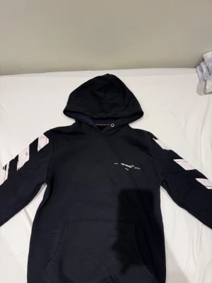 Svart Off-White hoodie med vita detaljer - Säljer en svart hoodie från Off-White med ikoniska vita pilar och streck på ärmar och rygg. Tröjan har huva, känguruficka och trycket 'SEEING THINGS' i blått nedtill på ryggen. Materialet är mjuk bomull och passformen är normal. Perfekt för dig som gillar streetwear och snygga detaljer.