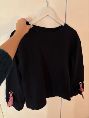 Svart tröja med rosa knyt från Vila - Unik långärmad tröja från Vila. Ärmarna har snygga rosa knytband och metallringar som detalj vid ärmsluten. Svart/marinblå