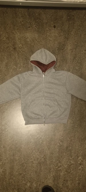 Grå hoodie med dragkedja - Grå hoodie med hel dragkedja och huva, insidan av huvan är brun. Tröjan har två fickor framtill och ribbade muddar vid ärmslut och nederkant.. Formen passar mig  som är (175cm 70kg) men den är ganska cropped och har baggy armar skulle även passa någon större.