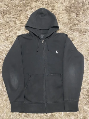 Svart hoodie från Polo Ralph Lauren - Svart hoodie från Polo Ralph Lauren med dragkedja och huva. Klassisk design med vit broderad logga på bröstet, snörning i huvan och två fickor framtill. Tillverkad i mjuk bomull och har långa ärmar. Perfekt för chill dagar och streetwear.