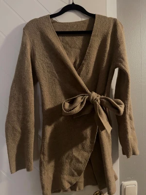 Beige omlottkofta med knytband - Säljer en beige omlottkofta med v-ringning och långa ärmar. Koftan har ett brett knytband i midjan 