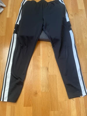 Svarta Adidas träningsbyxor med dragkedja - Svarta träningsbyxor från Adidas med vita och svarta ränder längs sidorna. Byxorna har dragkedja på ena sidan vid bensluten och klassisk Adidas-logga på benet. Knyten på insidan av midjan. 