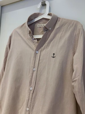 Beige skjorta från The Blue Uniform - Snygg beige skjorta från The Blue Uniform med klassisk krage och knappar framtill. På bröstet finns en broderad svart ankare som detalj. Skjortan är långärmad och tillverkad i mjuk bomull, perfekt för en chill och stilren look.