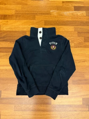 Mörkblå sweatshirt från Abercrombie & Fitch - Mörkblå sweatshirt från Abercrombie & Fitch med hög ribbad krage, tre knappar och broderad logga på bröstet. Tröjan har känguruficka framtill och är tillverkad i mjuk bomull. Perfekt för en chill och stilren look. Sweatshirten passar även en större M. 