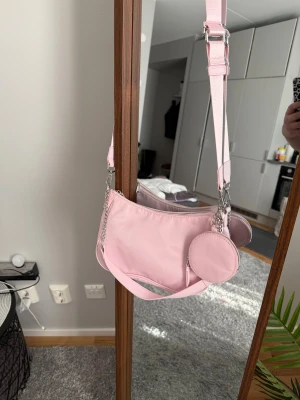 Ljusrosa axelväska med kedjedetalj - Snygg ljusrosa axelväska med justerbar rem och silverfärgad kedjedetalj. Väskan har en rund liten extra ficka framtill med dragkedja, perfekt för småsaker. Materialet är mjuk syntet och väskan stängs med dragkedja upptill. Enkel och trendig design.