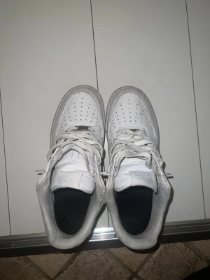Nike Air Force 1 vita sneakers - Säljer ett par klassiska Nike Air Force 1 sneakers i helvitt. Skorna har snörning, perforerad tå och platt sula. Märket syns tydligt på tungan och hälen. Perfekt för dig som gillar stilrena och tidlösa sneakers.