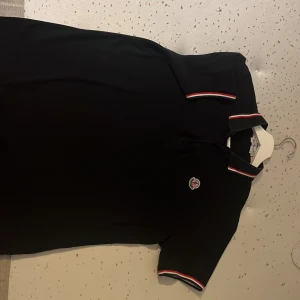 Svart Moncler pikétröja med röd/vit kant - Svart pikétröja från Moncler med klassisk krage och korta ärmar. Snygga rödvita detaljer på krage och ärmslut samt Moncler-logga broderad på bröstet. Perfekt för dig som gillar stilrena och exklusiva plagg med sportig vibe.
