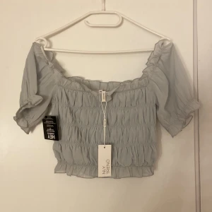 Ljusgrå croppad blus från NLY Trend - Supersöt ljusgrå blus från NLY Trend med croppad passform och offshoulder-design. Toppen har korta puffärmar, smockad struktur och volangdetaljer vid kanterna. Perfekt för dig som gillar romantisk och trendig stil.