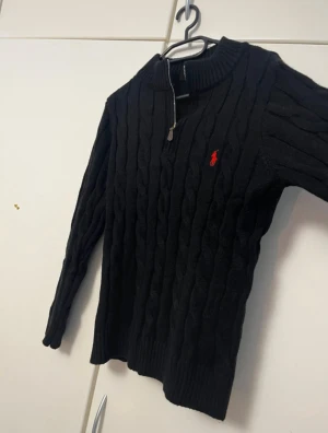 Polo Ralph Lauren tröja - Svart kabelstickad tröja från Polo Ralph Lauren med halv dragkedja och röd broderad logga på bröstet. Tröjan har ribbad krage, muddar och är långärmad. Perfekt för kyliga dagar och ger en klassisk, stilren look.