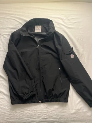 Svart vindjacka från Moncler - Svart vindjacka från Moncler med klassisk logga på ärmen och dragkedja framtill. Jackan har hög krage, elastiska muddar och fickor med dragkedja. Insidan har ett färgglatt serietryck. Perfekt för dig som gillar stilren och sportig look.