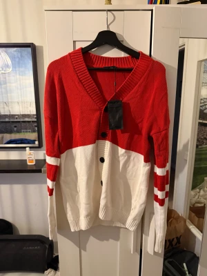 Röd och vit kofta Dorian Cardigan S (M) - Säljer en snygg kofta i rött och vitt från Dorian. Modellen har bred ribbad krage, knäppning framtill med svarta knappar och coola ränder på ärmarna. Tillverkad i bomull och passar perfekt för dig som gillar en sportig och avslappnad stil. Mer (Strl M)