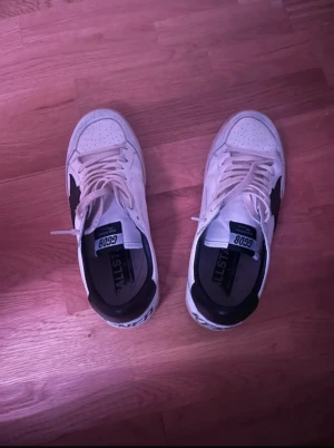 Golden Goose vita sneakers med svart detalj - Golden Goose sneakers i vitt skinn med svart stjärndetalj på sidan och svart häl. Platt sula i svart, snörning framtill och perforerad tå. Texten 'SNEAKERS' broderad på hälen. Kommer med originalkartong och dustbag.