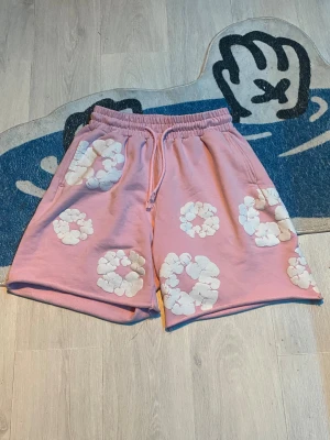 Rosa Denim Tears shorts med blommor - Säljer ett par rosa shorts från Denim Tears med vita blommiga tryck över hela plagget. De har elastisk midja med snörning och är tillverkade i mjuk bomull. Perfekta för en chill och trendig sommarstil.