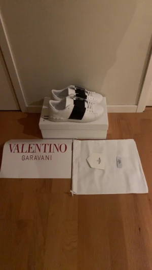 Valentino Garavani vita sneakers - Snygga vita sneakers från Valentino Garavani med bred svart rem över vristen och klassisk snörning. Skorna har rund tå, platt sula med mönstrad undersida och är tillverkade i skinn. Kommer med originalkartong och dustbag. 
