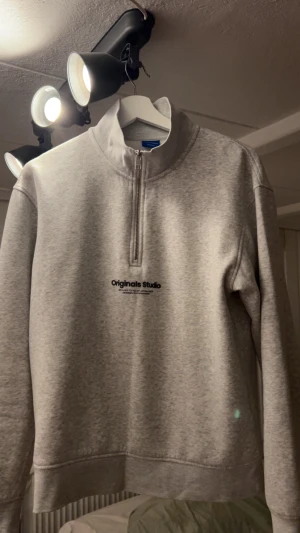 Grå half zip sweatshirt Jack & Jones - Snygg grå sweatshirt från Jack & Jones Originals Studio med half zip och hög krage. Tröjan har svart tryck på bröstet och är långärmad med en relaxed passform. Perfekt för chill dagar och enkel att matcha med jeans eller andra byxor