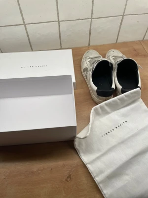 Oliver Cabell vita sneakers med grå detaljer - Snygga vita sneakers från Oliver Cabell med grå snörning och grått märke på sidan. Skorna har svart hälparti och är tillverkade i skinn. Klassisk rund tå och platt sula. Levereras med originalkartong och dustbag.