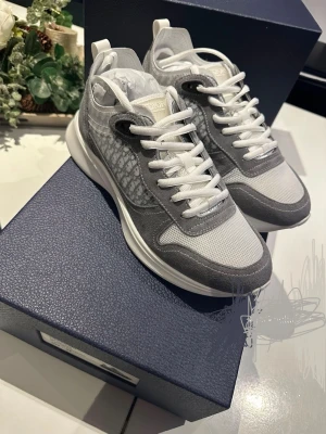  Grå/vita sneakers med monogram - Snygga sneakers i grått och vitt med klassiskt monogram på sidan. Skorna har vita snören, meshdetaljer och partier i mocka för en lyxig känsla. Sulan är vit och har rundade mönster undertill. Perfekta för dig som vill sticka ut med exklusiva detaljer.