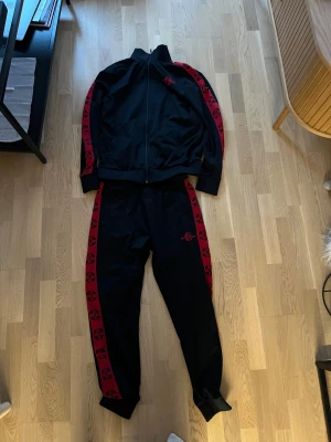 Svart Better Bodies set med röda detaljer - Svart set från Better Bodies i storlek M med röda logoband längs ärmarna och dragkedja framtill. Materialet är mjukt och syntetiskt, perfekt för träning. Långärmad modell med ribbade muddar och sportig look. Röd broderad logga på bröstet för extra stil. OBS byxorna saknar snöre, se bild 3.