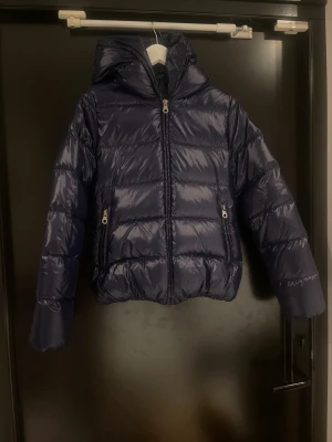 Mörkblå Duvetica pufferjacka med huva - Säljer en mörkblå pufferjacka från Duvetica med glansig yta, huva och två fickor med dragkedja. Jackan är fodrad med dun och har yttertyg i polyamid. Perfekt för kalla dagar och har en snygg, modern look.