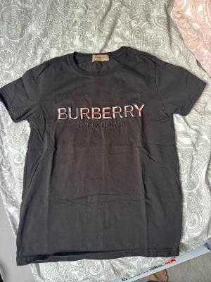 Svart Burberry t-shirt med broderad logga - Svart t-shirt från Burberry med broderad logga i vitt, rött och svart på bröstet. Klassisk rund halsringning och korta ärmar. Tillverkad i mjuk bomull för en skön känsla. Perfekt för dig som gillar stilrena och exklusiva plagg.